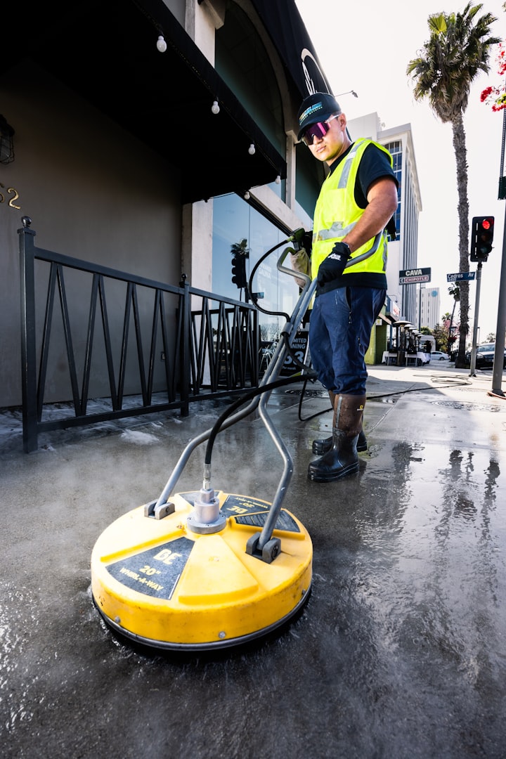A person in a gray cap, blue overalls, and yellow rubber boots is using a pressure washer on a driveway near a modern garage. The pressure washer is on the ground beside them, with a hose connected. There are potted plants nearby and wet spots on the pavement.